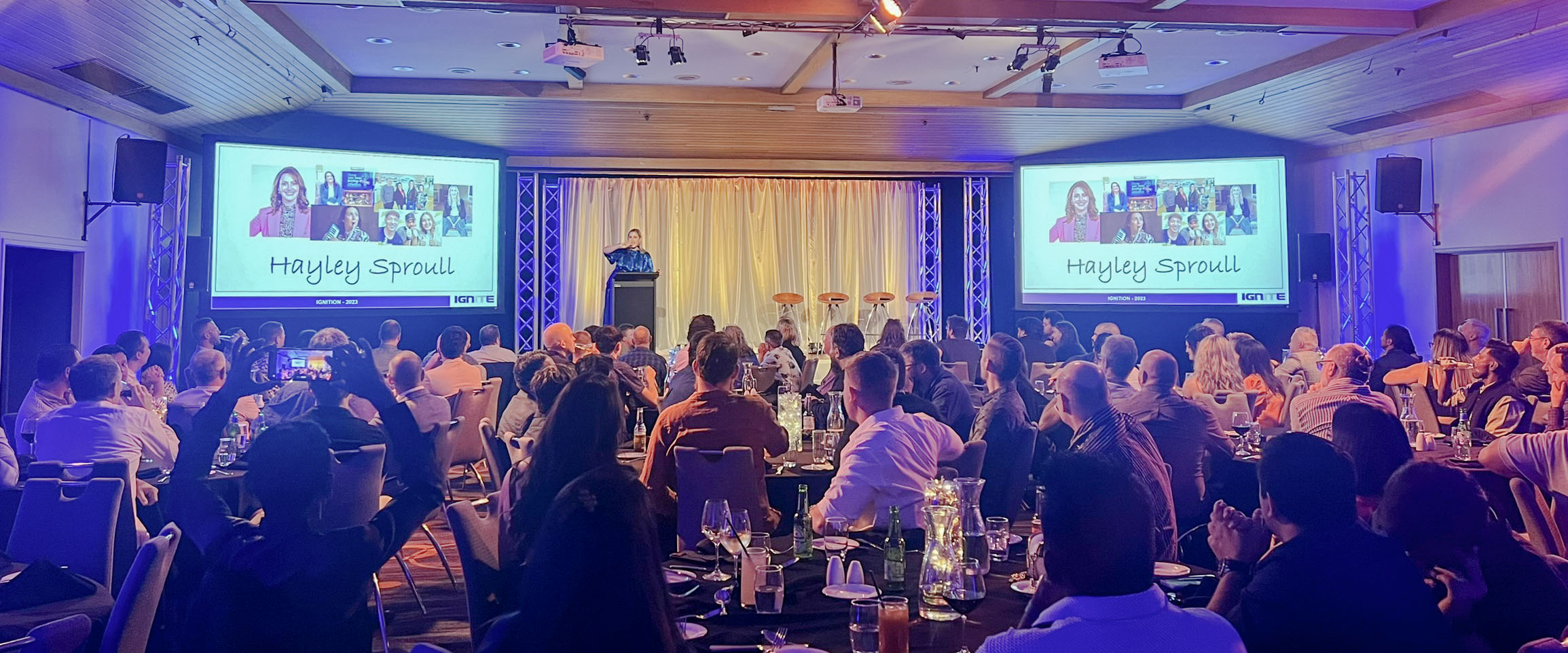 Conference presentation with two large screens, stage lighting and a seated audience